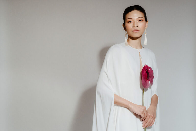 A model in a photo studio is dressed in a modern wedding dress and cape. She hold a single flower stem and the image illustrates an article on the best AI photo editing tools for wedding photographers.