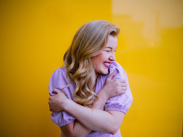 A Scandinavian Blonde girl in a lilac dress stands in front of a yellow wall with her arms hugging herself for an article on protecting your wedding photography business