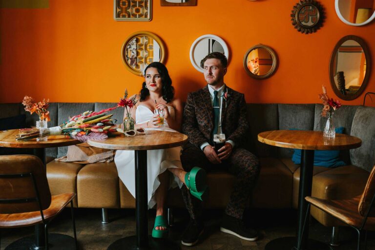 A bride and groom sitting at a cafe table, posing for a portrait. The background shows a retro cafe with eclectic decor. This image could be used to illustrate testimonials for wedding photographers, showcasing the happy couple.
