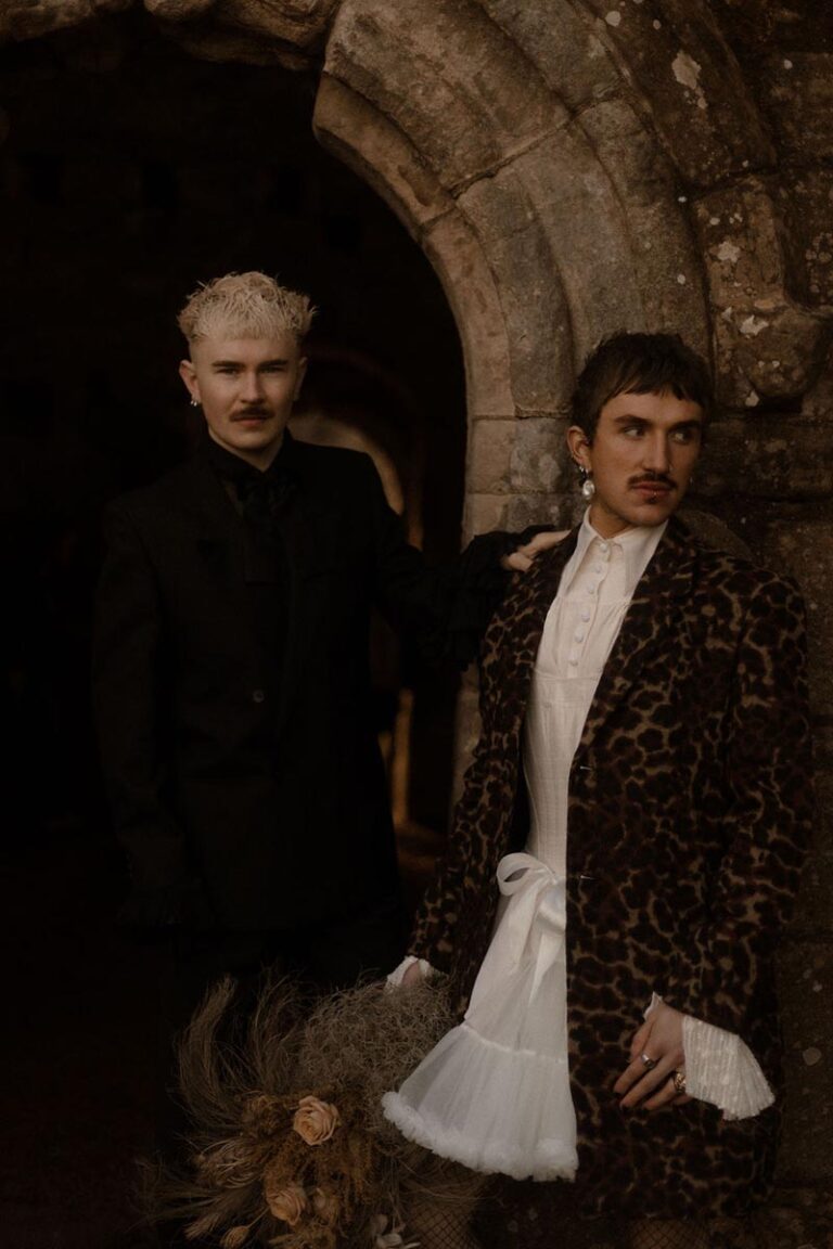 A couple model as two grooms for the styled shoot part of photography farm's thrive conference. They stand outside an old church dressed in wedding outfits. This was taken at our thrive conference by Esme Whiteside.