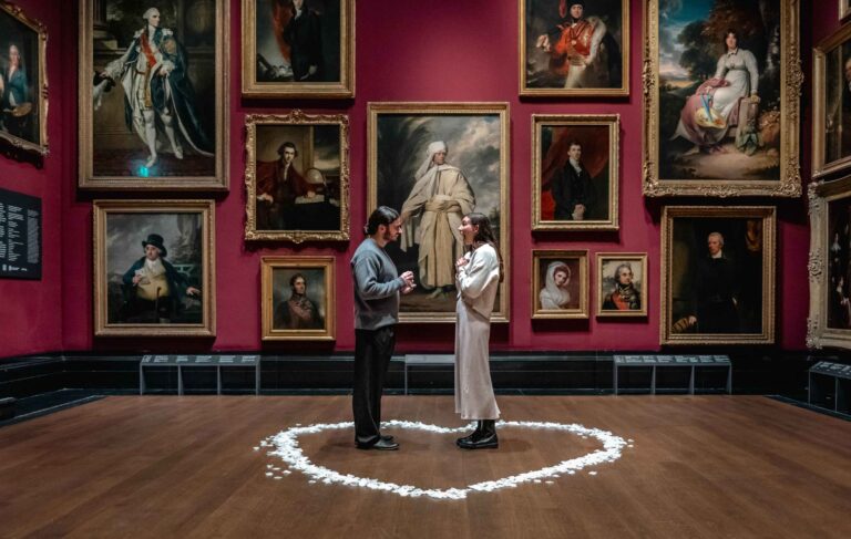 A couple stand in a circle of petals in front of a gallery wall of paintings for Proposal Photography at the National Portrait Gallery. The girl has a surprised expression while the man holds an engagement ring.