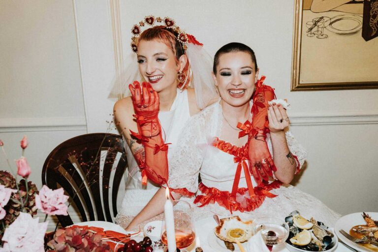Two brides dressed in playful and cool wedding attire pose joyfully at a lavishly set table, showcasing unconventional wedding photography styles