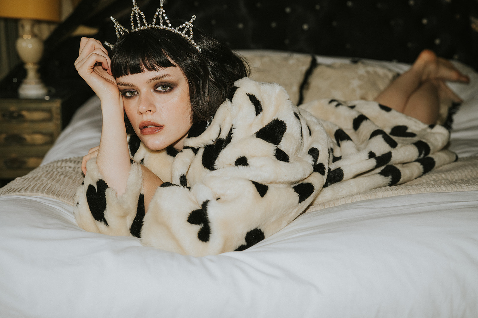 A woman wearing a spotted fur coat and a crown lies on a bed, gazing at the camera representing a styled photoshoot concept related to wedding photography cancellations.