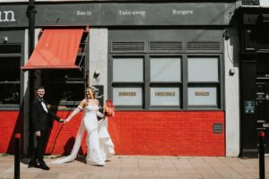 A bride and groom walk hand in hand, playfully. A great image to go with a blog about A bride and groom share a kiss beside a vintage red double-decker bus decorated for their wedding day — a joyful moment illustrating The Offboarding Process For Wedding Photographers.