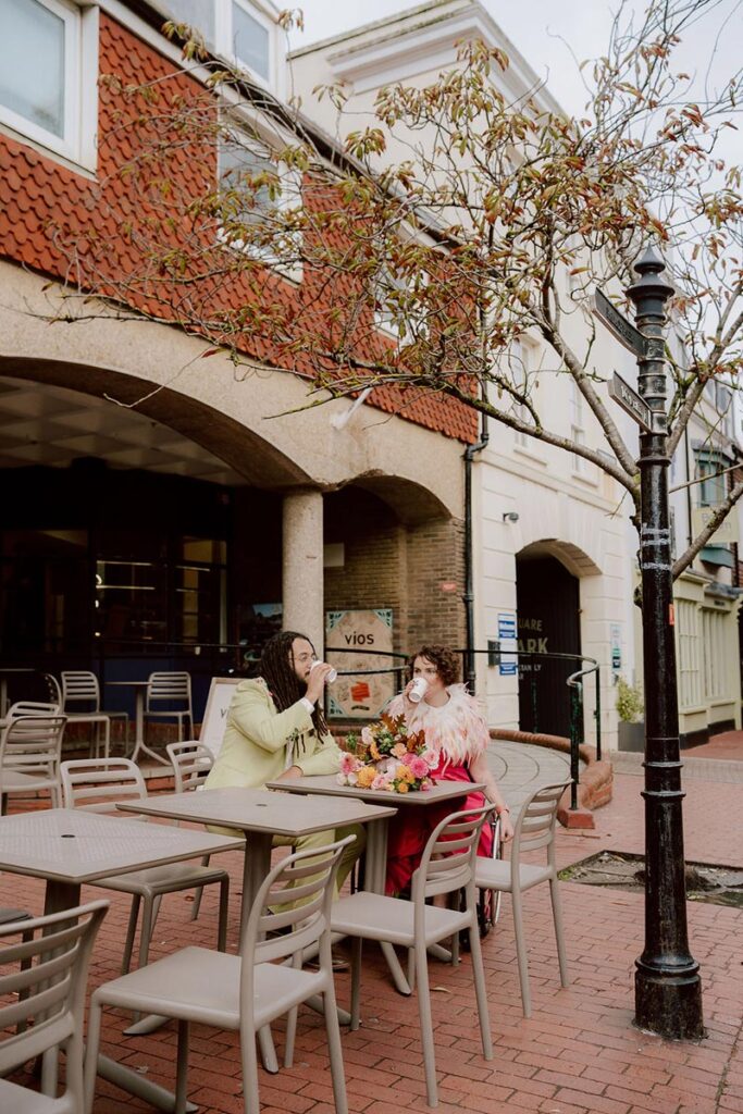 Brighton wedding photographer Kim Williams’ Thrive shoot couple Meg and Xander sharing coffee and laughter at an outdoor café, styled in bold pink and embroidered green suits.